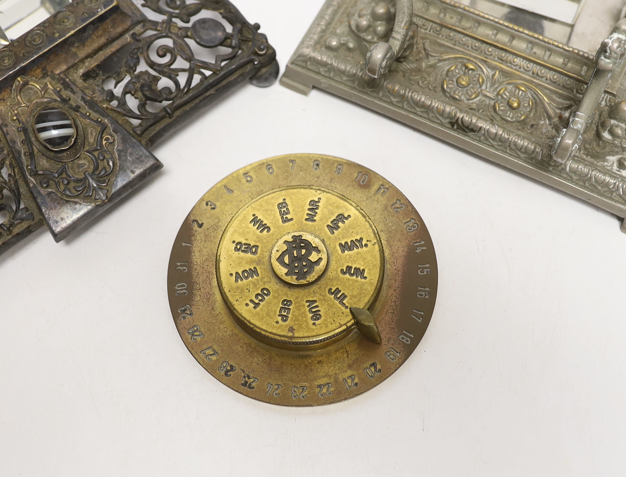 Three cast metal and cut glass ink stands, including a bronze inkwell in the form of the head of Bacus, and a brass calendar display from the Banca Commerciale Italiana, London Branch, tallest 13cm